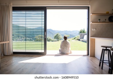 Rear view of young woman sitting by patio door at home. Copy space.