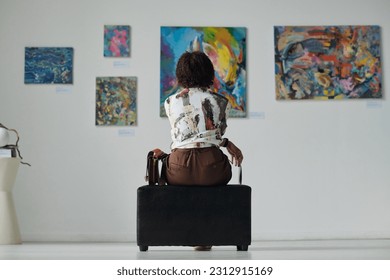 Rear view of young woman sitting on couch and looking at paintings on the wall, she enjoying art in gallery