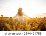 A rear view of a young woman in a straw hat walking in a golden wheat field. A beautiful woman enjoys the sunset and feels free. The concept of relaxation, freedom, walks.