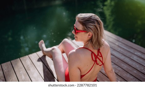 Rear view of young woman in swimsuit sitting on pier by lake during summer vacation in mountains. - Powered by Shutterstock