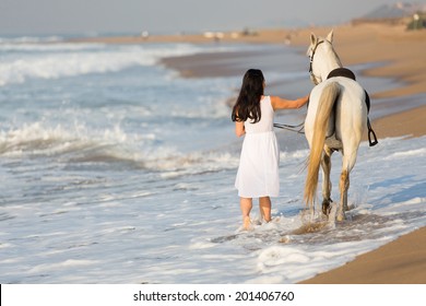 Rear View Of Young Woman Walking A White Horse On Beach