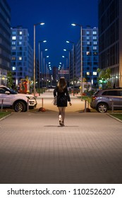 Rear View Of Young Woman Wearing Black Coat Walking Down The Street, Street Lights Towards Her. A Female With Beautiful Legs And Long Hair Goes Along The Night City Sidewalk.