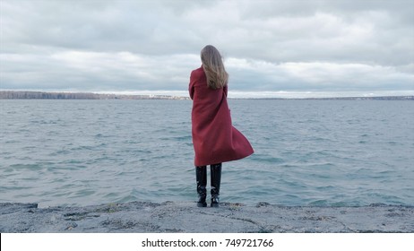 Rear view of a young woman wearing a red coat against a bright blue sky and sea on a holiday beach, outdoors. Rear view of alone young adult girl standing on the beach, looking to sea. Travel and