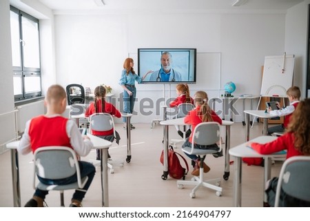 Rear view,class of pupils sitting, listening to teacher and wathing online lecture with doctor in classroom