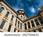 Rear views of the Illinois State Capitol Building in Springfield, Illinois, USA. Cloudy blue skies overhead. The US flag, State of Illinois Flag, and Black History Month Flag fly atop the dome.