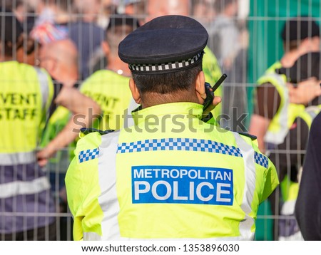 Rear Waist Up View of Male Metropolitan Police Officer in Yellow Flourescent Jacket Listening to His Radio.  Event Stewards and a Crowd Behind