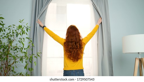 Rear of young woman with long red curly hair standing in living room at window and opening curtains in the morning. Back view.