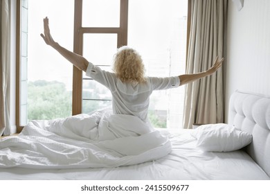 Rearview 60s female welcoming new happy day, spread arms seated on comfortable bed, stretching body after healthy night sleep in cozy bedroom at home or hotel on vacation. Good morning, quality rest - Powered by Shutterstock