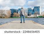 Rearview of gray haired businessman in warm jacket and scarf standing on river bridge, looking at office buildings. Moving new apartment, build new city area and site development. Modern urban area