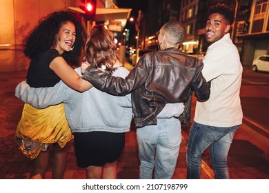 Rearview Of A Group Of Friends Walking In The City At Night. Group Of Vibrant Young People Laughing Joyfully While Embracing Each Other. Multicultural Friends Going Out Together On The Weekend.