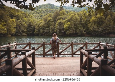 rearview landscape with Caucasian blonde woman wearing casual clothes relaxing and enjoying the view on wooden dock by a lake surrounded by green forest in Yedigoller National Park Turkey