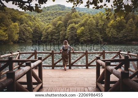 rearview landscape with Caucasian blonde woman wearing casual clothes relaxing and enjoying the view on wooden dock by a lake surrounded by green forest in Yedigoller National Park Turkey