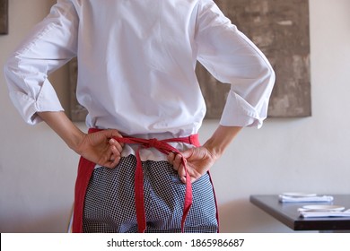 Rearview of a midsection of a female chef standing in a restaurant and tying apron strings with a painting hanging on the wall in the background. - Powered by Shutterstock