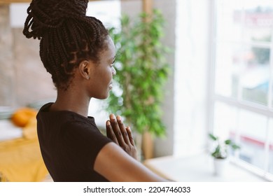 Rearview mindful and meditating young multicultural woman in black sportswear practicing yoga exercises, standing and keeping hands in namaste mudra position. Fitness at home, looking out window