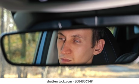 Rearview mirror reflection of dark-haired man sitting at steering wheel upset because of new car breakdown standing on forest roadside in autumn - Powered by Shutterstock