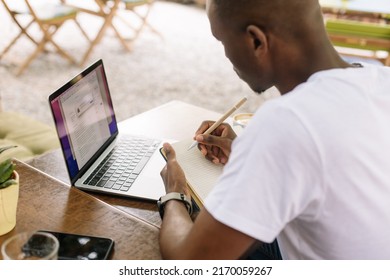 Rearview Multicultural Dark Skin Man Using Laptop, Taking Notes In Workbook Paper During Internship Study In Street Internet Cafe Outdoor. Businessman Writing Business Plan, Annual Report. High Angle