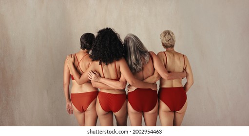 Rearview Shot Of Four Women Of Different Ages Embracing Their Natural And Aging Bodies. Group Of Women Embracing Each Other While Wearing Red Underwear And Standing Together In A Studio.