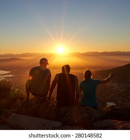 Rearview Silhouettes Of Three Friends Sitting Together On A Nature Trail Watching A Golden Sunrise