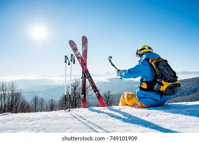 Rearview view of a skier with backpack and ski equipment sitting on the snow on top of the mountain taking selfies with his camera on a selfie stick while resting after skiing at the winter resort