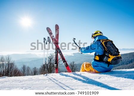 Rearview view of a skier with backpack and ski equipment sitting on the snow on top of the mountain taking selfies with his camera on a selfie stick while resting after skiing at the winter resort