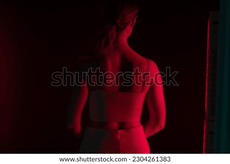 Rearview of a woman receiving red light therapy on her back. Woman standing next to a red light device in a cosmetic spa. Anti-aging skincare treatment.