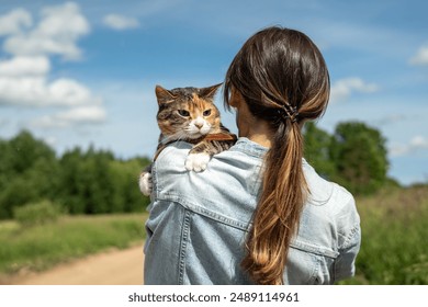 Rearview of woman strolling down with tabby cat in collar, perched on shoulder on sunny day in park outdoors. Pet owner caring about kitty and spending time together on summer day - Powered by Shutterstock