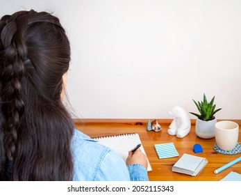 Rearview Of A Woman Writing A Letter In A Notebook With Copy Space Wall. Girl Taking Notes On A Desk With Plant And Sticky Notes. Planning On Notepad With A Coffee Cup. Home Office For Studying