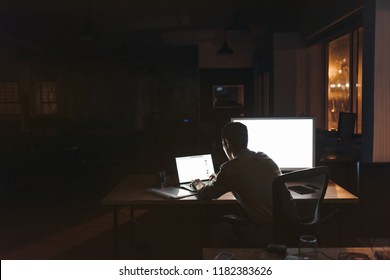Rearview of a young businessman sitting alone at a desk in a dark office working on a laptop late in the evening