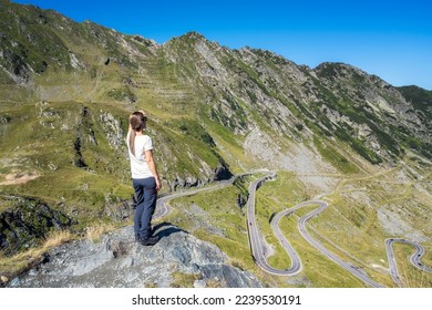 Rearview of a young woman standing at a cliff edge looking from the top at famous Transfagarasan mountain road in Romania.	Beautiful girl enjoying the nature beauty. 