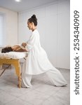 Rebalancing process. Beautiful masseuse in white outfit makes body massage to lying on back woman in pro salon, vertical image, selective focus.