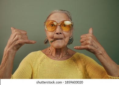 Rebel Serious Senior Woman Showing Shaka Sign, Smoking And Looking. Isolated Over Green Background. Hand Rolled Cigarettes. Vacation Concept. Casual Clothes. Gray Straight Hair. Orange Sunglasses