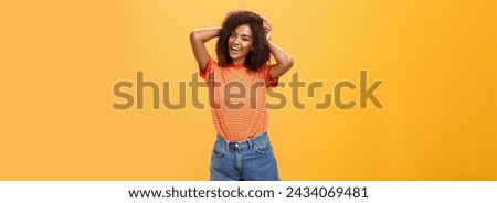 Rebellious playful and joyful african american girl with curly hairstyle making devil horns on head with index fingers smiling broadly showing daredevil character and charisma over orange wall