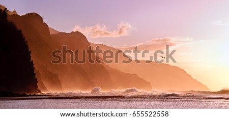Receding headlands of the Na Pali Kauai coastline illuminated at sunset over a stormy sea with a distant bird. The mountains off Ke'e beach are the start of the Kalalau Trail