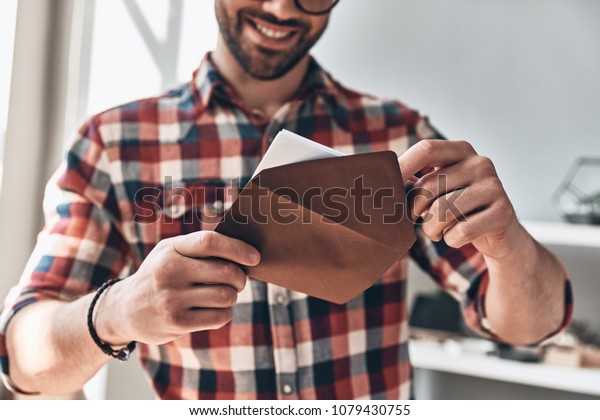 Receiving greeting card. Close up\
of young man opening envelope and smiling while standing\
indoors