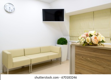 Reception And TV In Waiting Room Of Hospital Or Office. Light Clean Interior Of Clinic. Sofa In Entrance Area, Hall With Minimalist Design. Inside Modern Hallway With Nice Decor. Focus On  Flowers. 