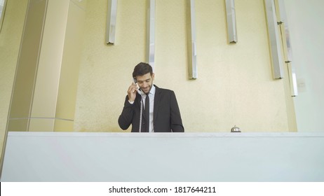 A Receptionist Or Concierge Man With Uniform At Front Desk Counter In Lobby Of Hotel Talking On The Phone With Guest About The Service Room. The Staff Employee Working.