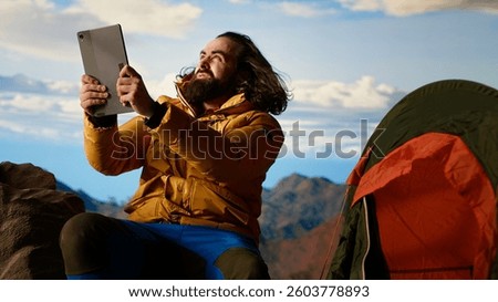 Reckless explorer struggling with wind and weak signal keep internet connection, camping on a scenic rocky mountain peak. Hiker in danger with extreme cold weather, foolish. Camera B.