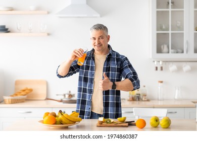 I recommend healthy food. Happy senior man drinking fresh juice, standing near table full of various fruits and showing thumb up in kitchen interior, empty space