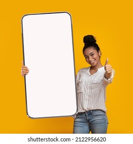 I Recommend. Portrait Of Smiling Black Female Teenager Holding Big Giant Cell Phone With White Blank Device Screen In Hand, Showing Thumb Up Gesture. Gadget With Empty Free Space For Mock Up, Banner