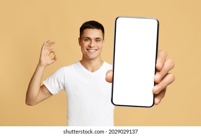 I Recommend. Portrait of smiling man holding smartphone with white blank device screen in hand, showing okay sign gesture, beige studio. Gadget with empty free space for mock up, isolated display - Shutterstock ID 2029029137