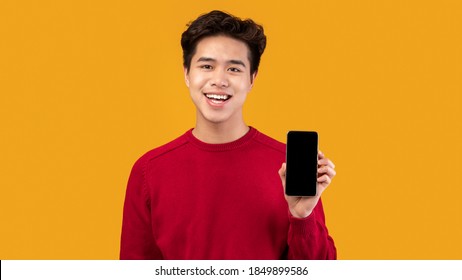 I Recommend This App. Smiling Asian Guy Holding Smartphone With Black Empty Screen In Hand And Showing It To Camera. Gadget With Blank Space For Mock Up, Isolated Over Orange Studio Background