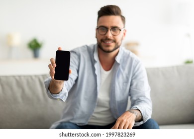 I Recommend This Website. Young Man Showing Smartphone With Blank Screen To Camera, Mockup For Your Ad Or Design. Guy Sitting On Sofa At Home, Holding Cellphone In Hand, Selective Focus