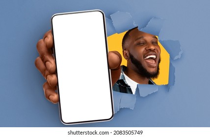 Recommendation. Excited black guy holding big smartphone with white blank screen in hand, showing close to camera through torn blue paper hole. Gadget with empty free space for mock up, banner