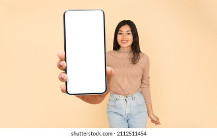 Recommendation. Portrait Of Excited Asian Lady Holding Big Smartphone With White Blank Screen In Hand, Showing Device Close Up To Camera. Gadget With Empty Free Space For Mock Up, Studio Banner