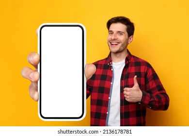 Recommendation. Portrait of excited guy holding big giant cellphone with white blank screen in hand, showing making thumb up gesture. Gadget with empty free copy space for mock up banner, yellow wall - Shutterstock ID 2136178781