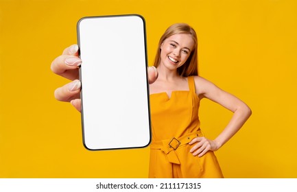 Recommendation. Portrait Of Excited Young Lady Holding Big Smartphone With White Blank Screen In Hand, Showing Device Close Up To Camera. Gadget With Empty Free Space For Mock Up, Yellow Orange Wall