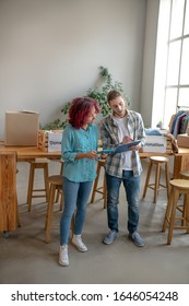 Reconciliation. Young Girl In A Blue Shirt With A Tablet And A Man In A Plaid Shirt With Copybook Standing Near The Table And Checking Data, Serious And Attentive.