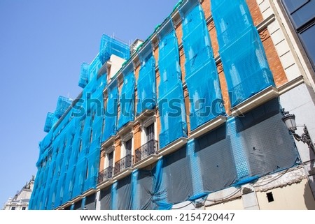 Reconstruction of facade of a historic classical apartment building on a city street in sunny day against sky. Blue facade construction mesh covers an old red house closed for renovation wall exterior