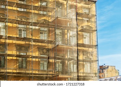 Reconstruction Of An Old Residential Building Covered With Green Protective Polypropylene Mesh. Scaffolding Near The Facade Of The Building Against The Blue Sky