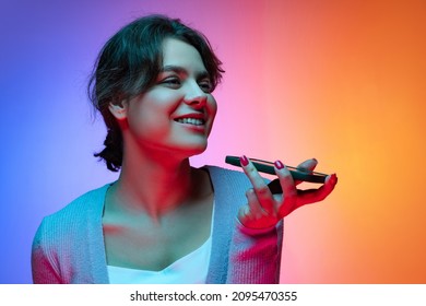 Recording voice message. Happy young pretty girl in warm gray cardigan with phone isolated on gradient blue orange neon background. Concept of emotions, facial expression, youth, aspiration. - Powered by Shutterstock